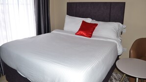 Ein Hotelzimmer mit einem großen Bett, einem roten Kissen, einem kleinen runden Tisch und einem Fenster mit Vorhängen.