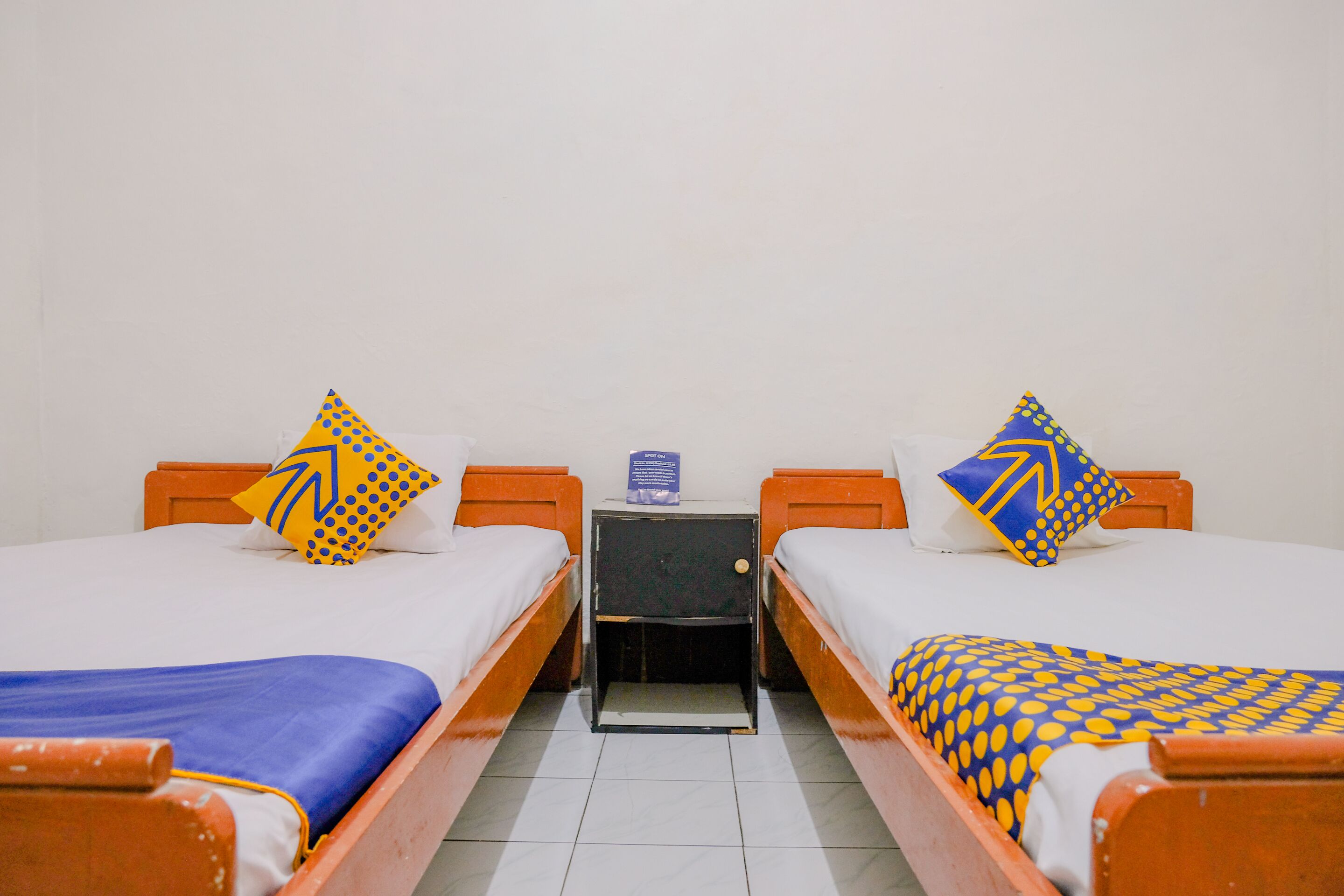 Een kamer met twee eenpersoonsbedden, elk voorzien van een gekleurd kussen en een blauwe sprei. Tussen de bedden staat een klein nachtkastje met een blauw bordje erop.