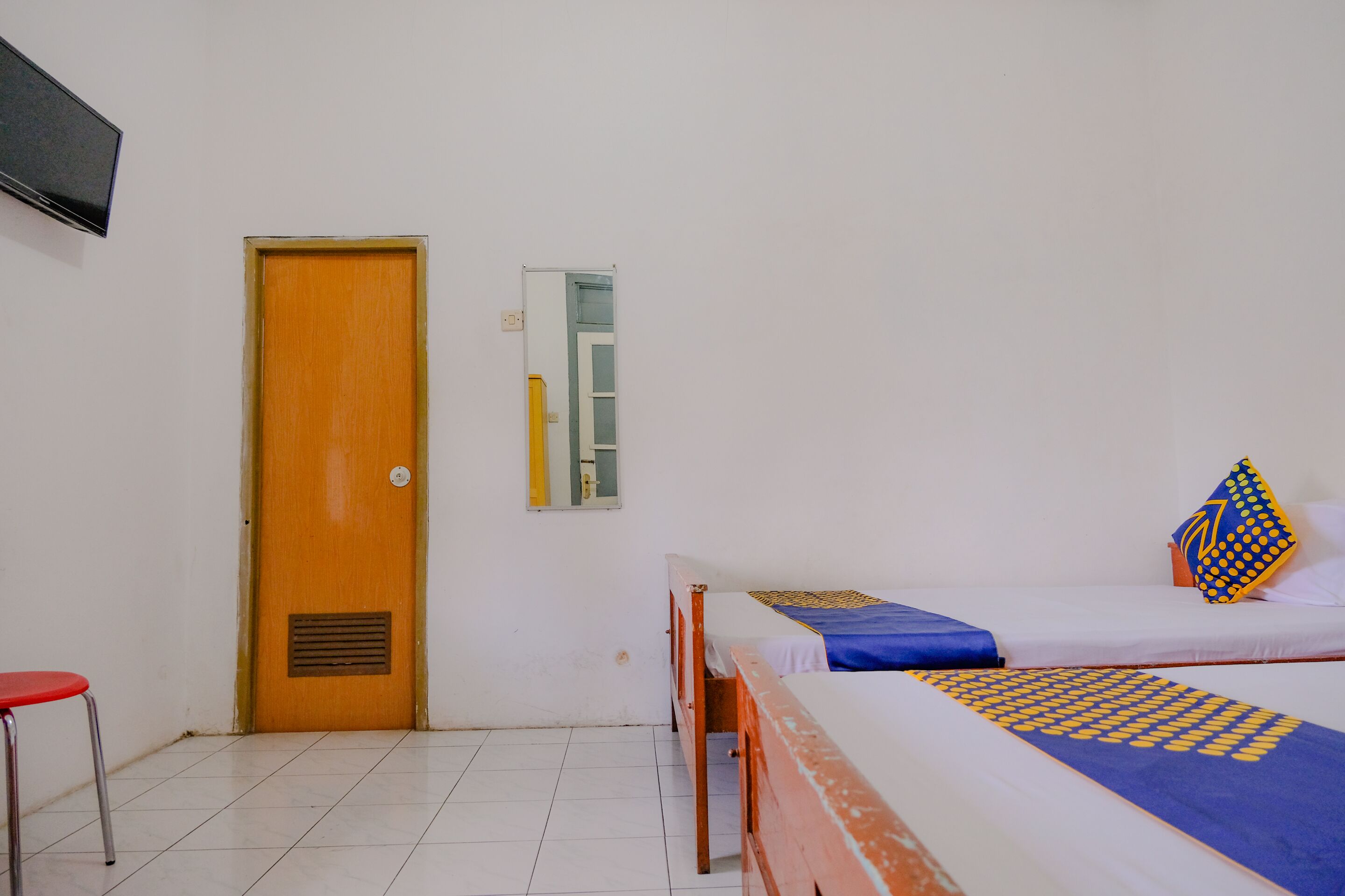 A room with two beds, a wooden door, a small stool, and a mirror on the wall.