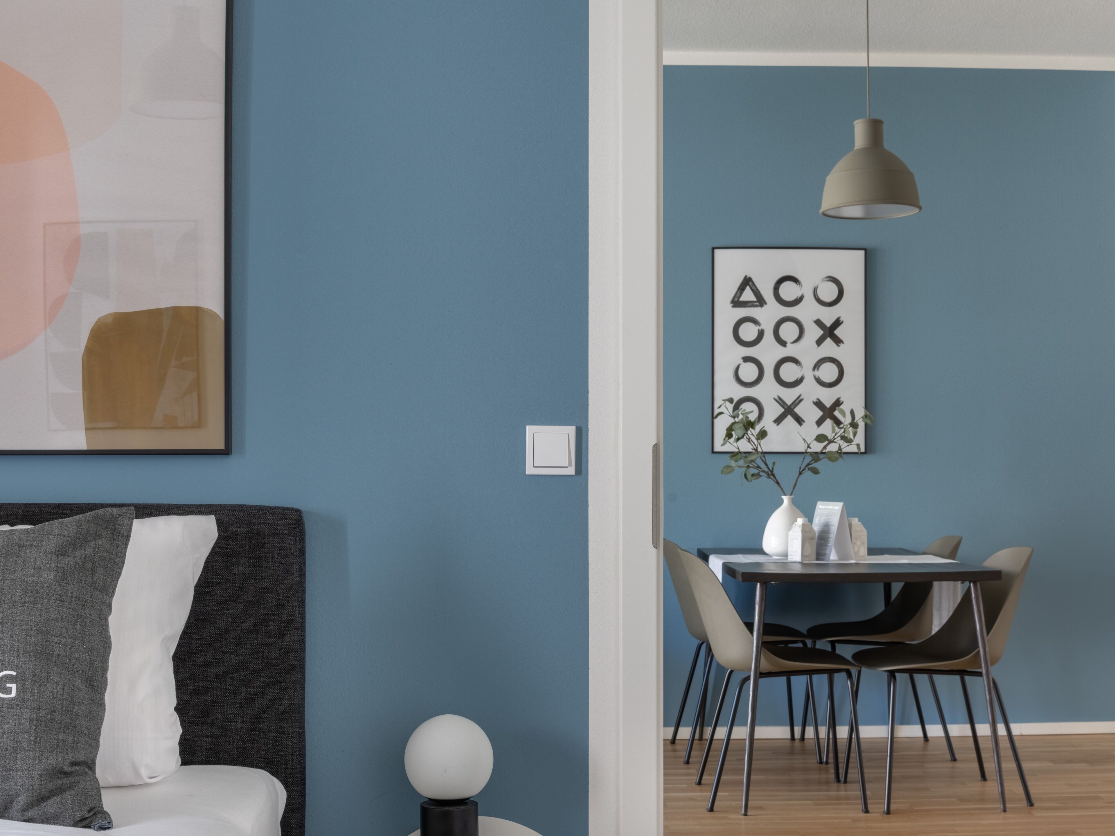 Quarto moderno com parede azul escura, sofá cinza com almofada branca, mesa redonda branca com cadeiras, luminária pendente e quadro com formas geométricas.