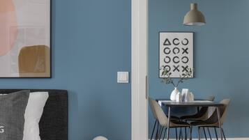 Une pièce moderne avec un mur bleu foncé, un canapé gris agrémenté d’un coussin blanc, une table ronde blanche avec des chaises, un luminaire suspendu et un tableau encadré aux motifs géométriques.