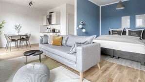 Un salon moderne avec un canapé gris, un pouf rond, une table à manger avec des chaises, un coin cuisine et un lit avec une tête de lit grise.