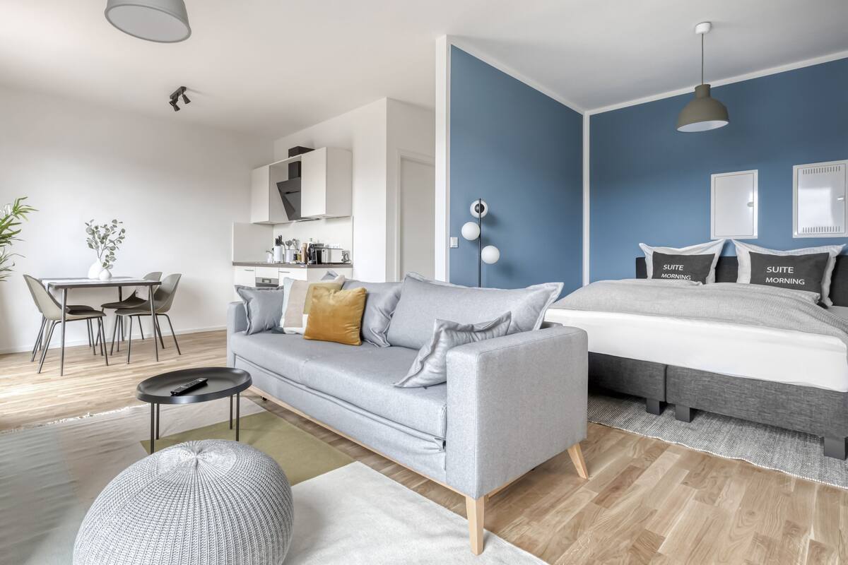 Een moderne woonkamer met een grijze bank, een rond hockertje, een eettafel met stoelen, een keukenhoek en een bed met een grijs hoofdeinde.