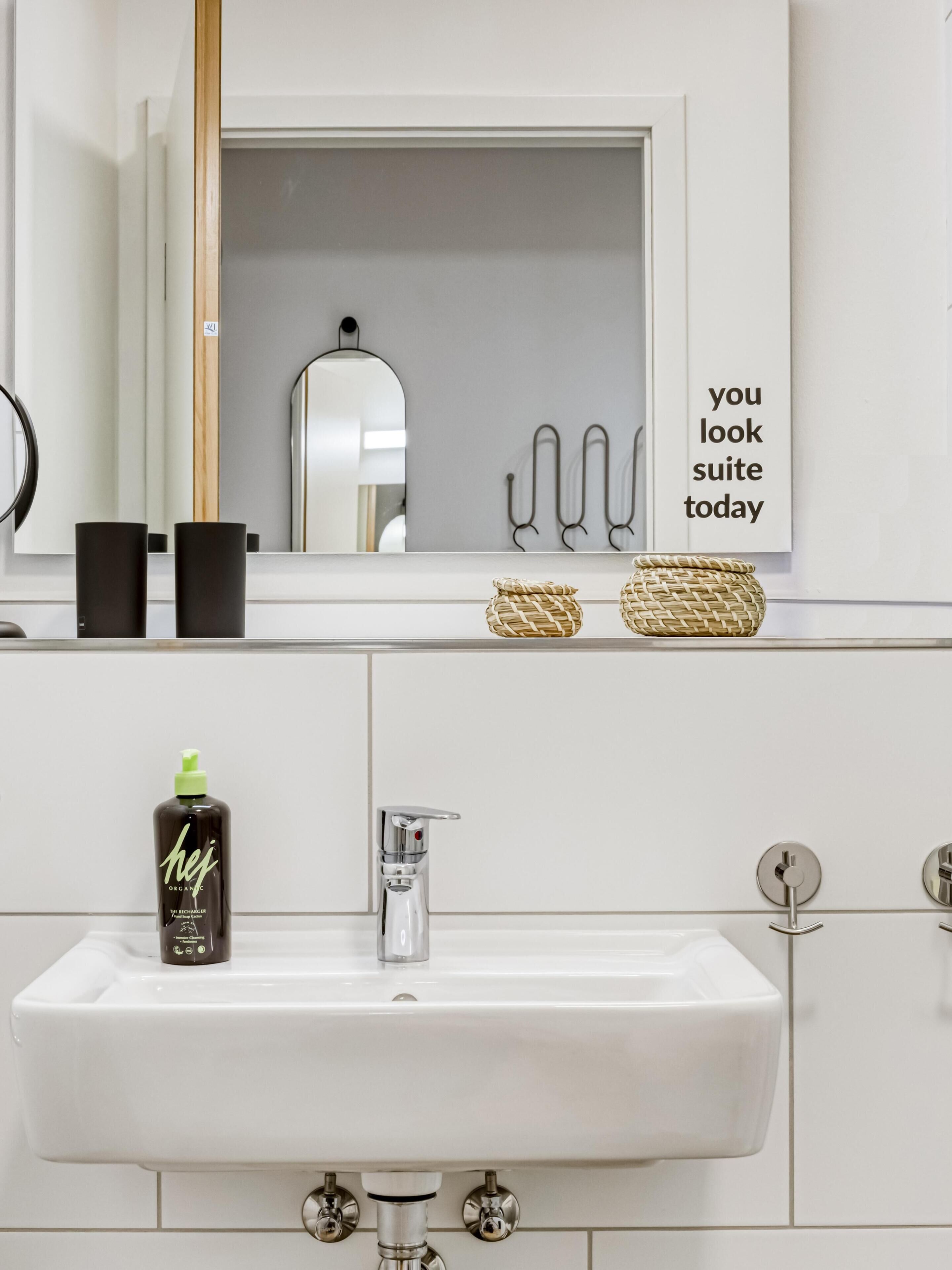 Une salle de bain avec un lavabo blanc, un miroir et une étagère sur laquelle sont posées deux tasses noires et deux paniers en osier.