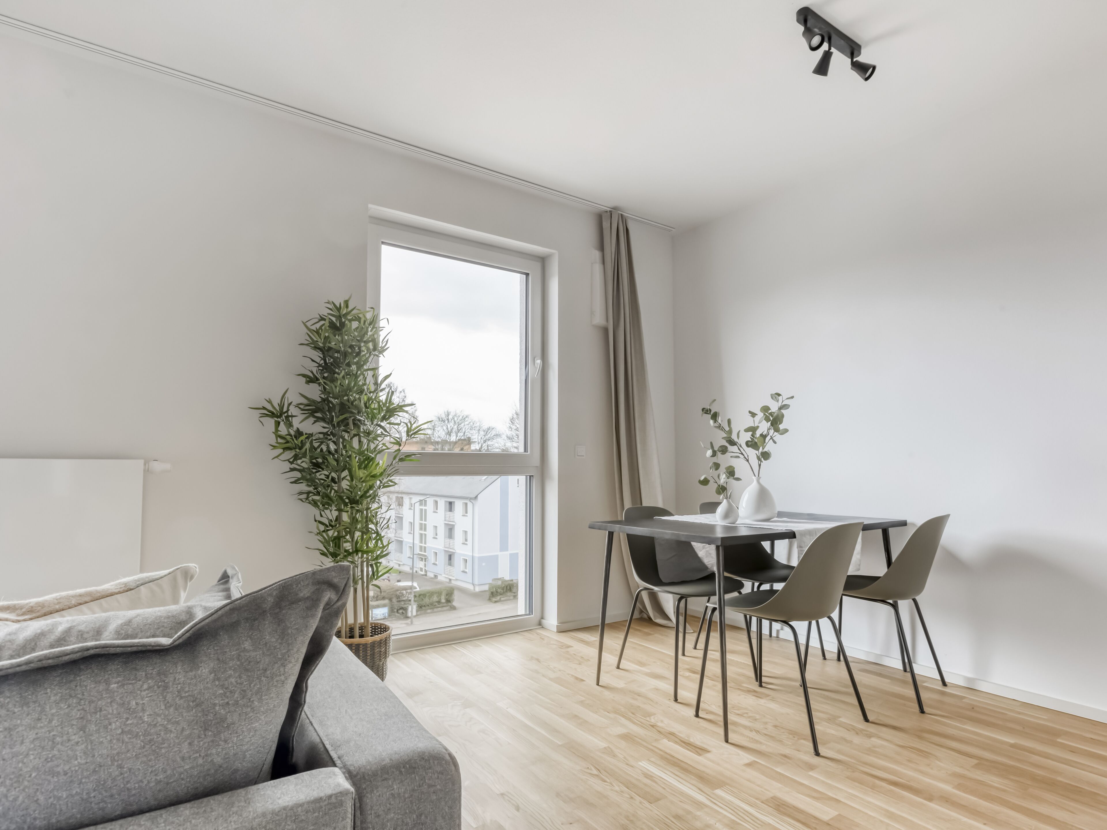 Un salon moderne avec un canapé gris, une table à manger avec des chaises, une plante en pot et une vue sur les immeubles à travers une grande fenêtre.