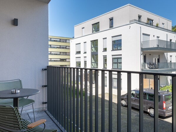 Un balcón con mesa y sillas, vista de un edificio con múltiples ventanas y balcones, y un coche aparcado al fondo.