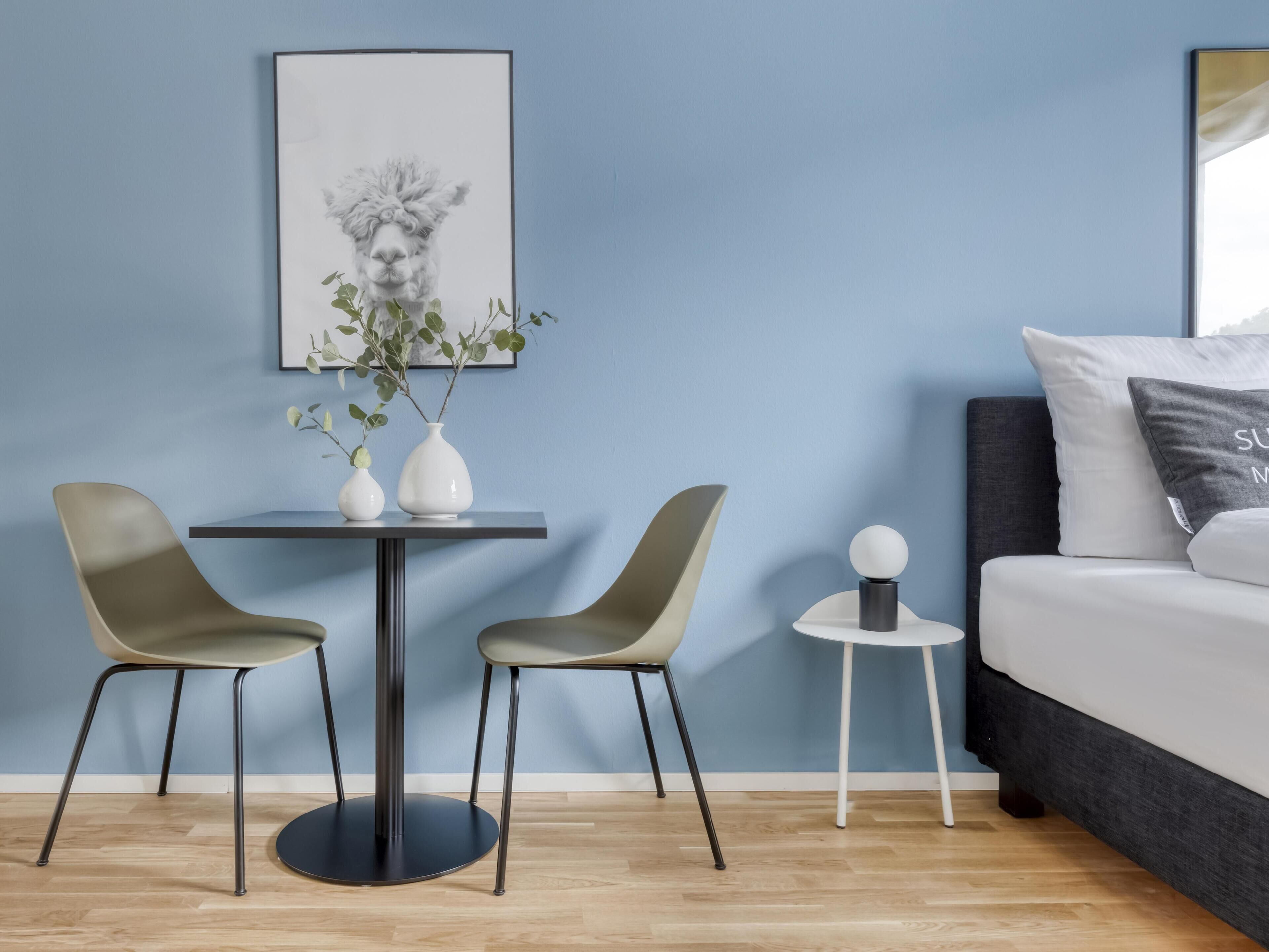 Una habitación moderna con una pared azul, una mesa redonda con dos sillas, una mesita auxiliar con una esfera decorativa, una cama con cabecero oscuro y un cuadro enmarcado sobre la mesa.