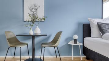 Una habitación moderna con una pared azul, una mesa redonda con dos sillas, una mesita auxiliar con una esfera decorativa, una cama con cabecero oscuro y un cuadro enmarcado sobre la mesa.