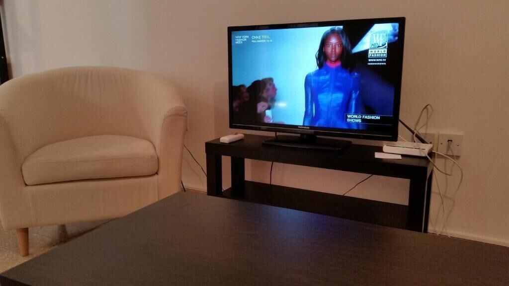 Een televisie op een zwarte standaard in een kamer met een beige fauteuil.