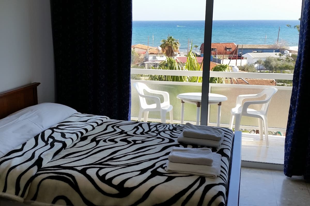 A hotel room with a bed, a view of the sea, and a balcony with chairs.