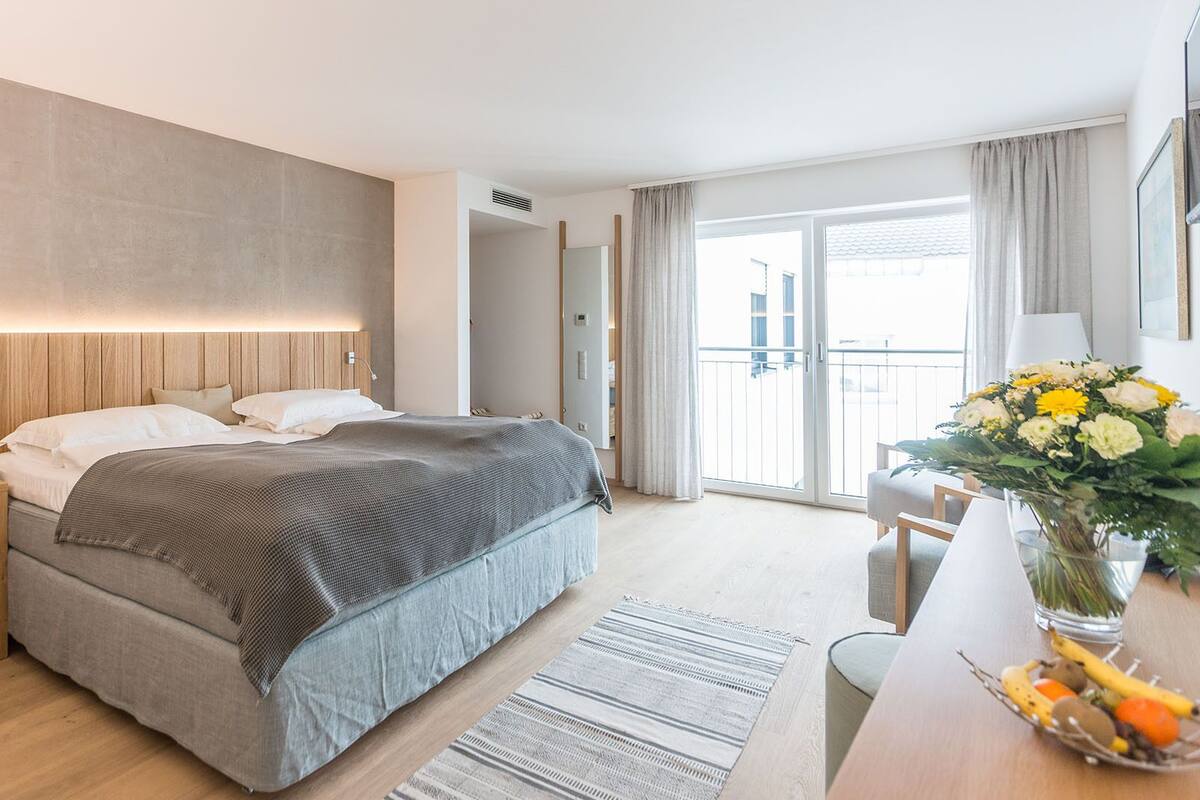 Quarto de hotel moderno com uma cama grande, cabeceira de madeira, mesas de cabeceira, uma escrivaninha com um vaso de flores e vista para o exterior por uma porta deslizante.