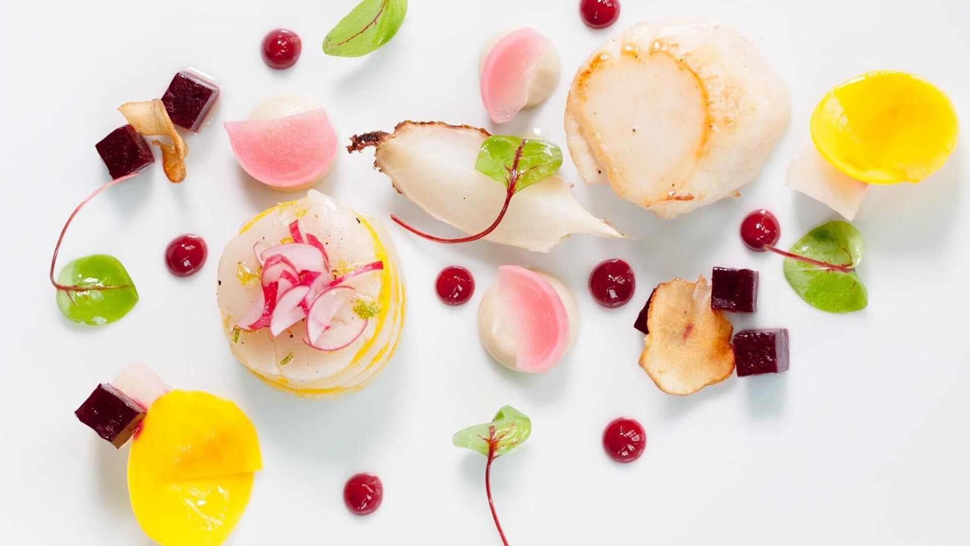 A variety of food items including scallops, radishes, and possibly mango slices, arranged on a white surface.