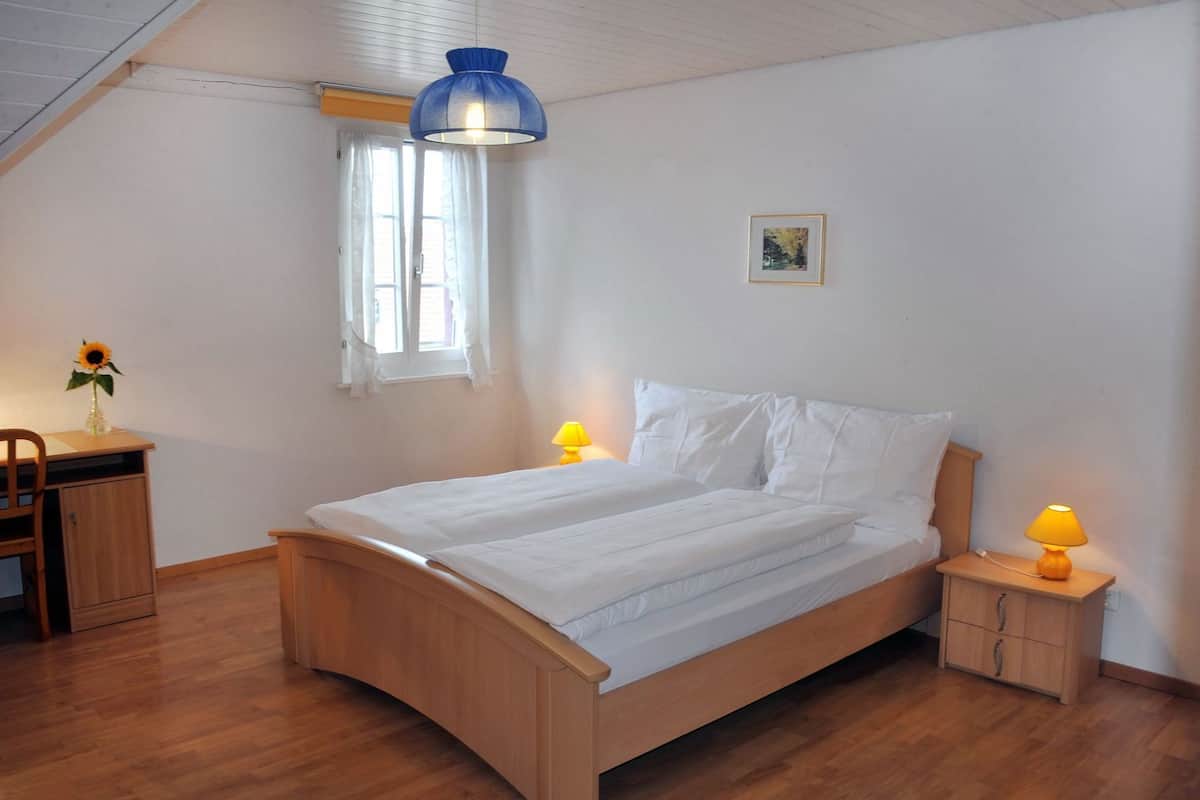 Un dormitorio con una cama de madera, dos lámparas de noche, una ventana con cortinas, un escritorio con una silla y un cuadro en la pared.