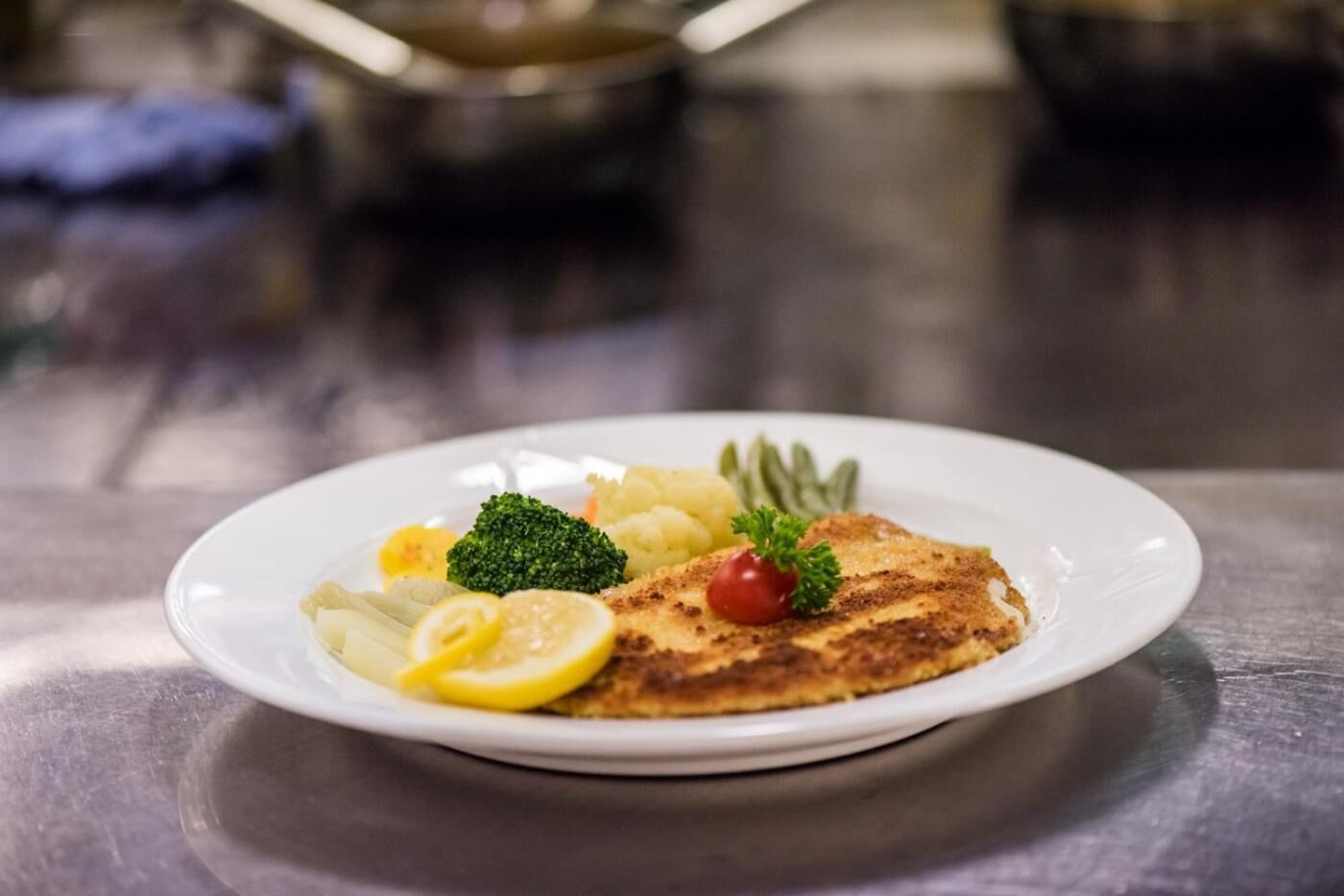 Une assiette contenant une escalope panée, des tranches de citron, des brocolis et une tomate cerise.