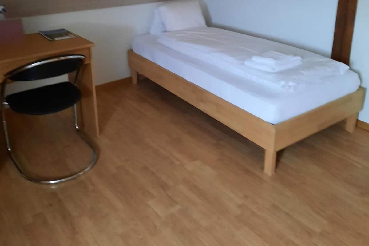 Una habitación pequeña con cama individual, techo inclinado, escritorio de madera y una silla.