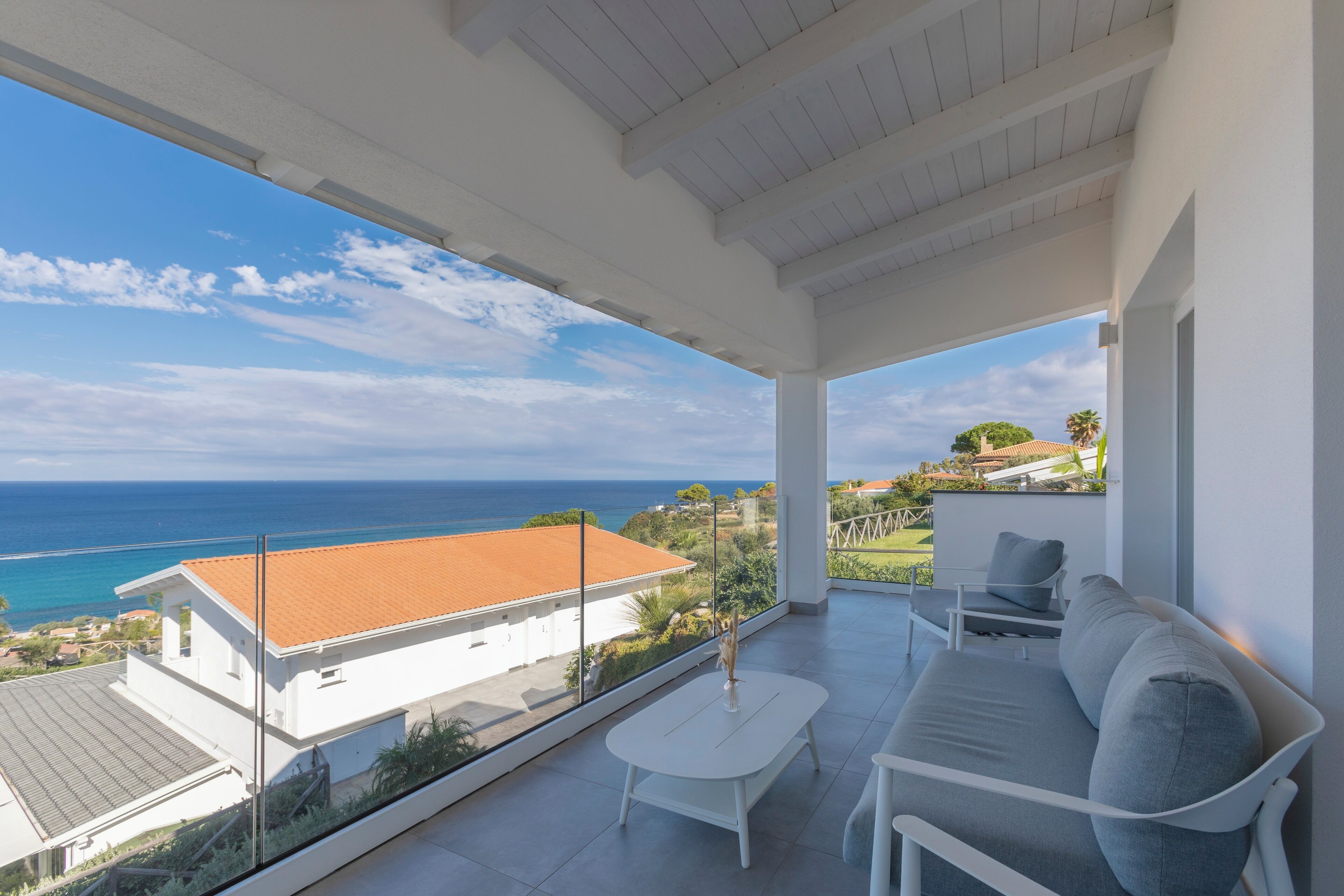 A balcony with a white sofa and table, overlooking a coastal view with a building and greenery.