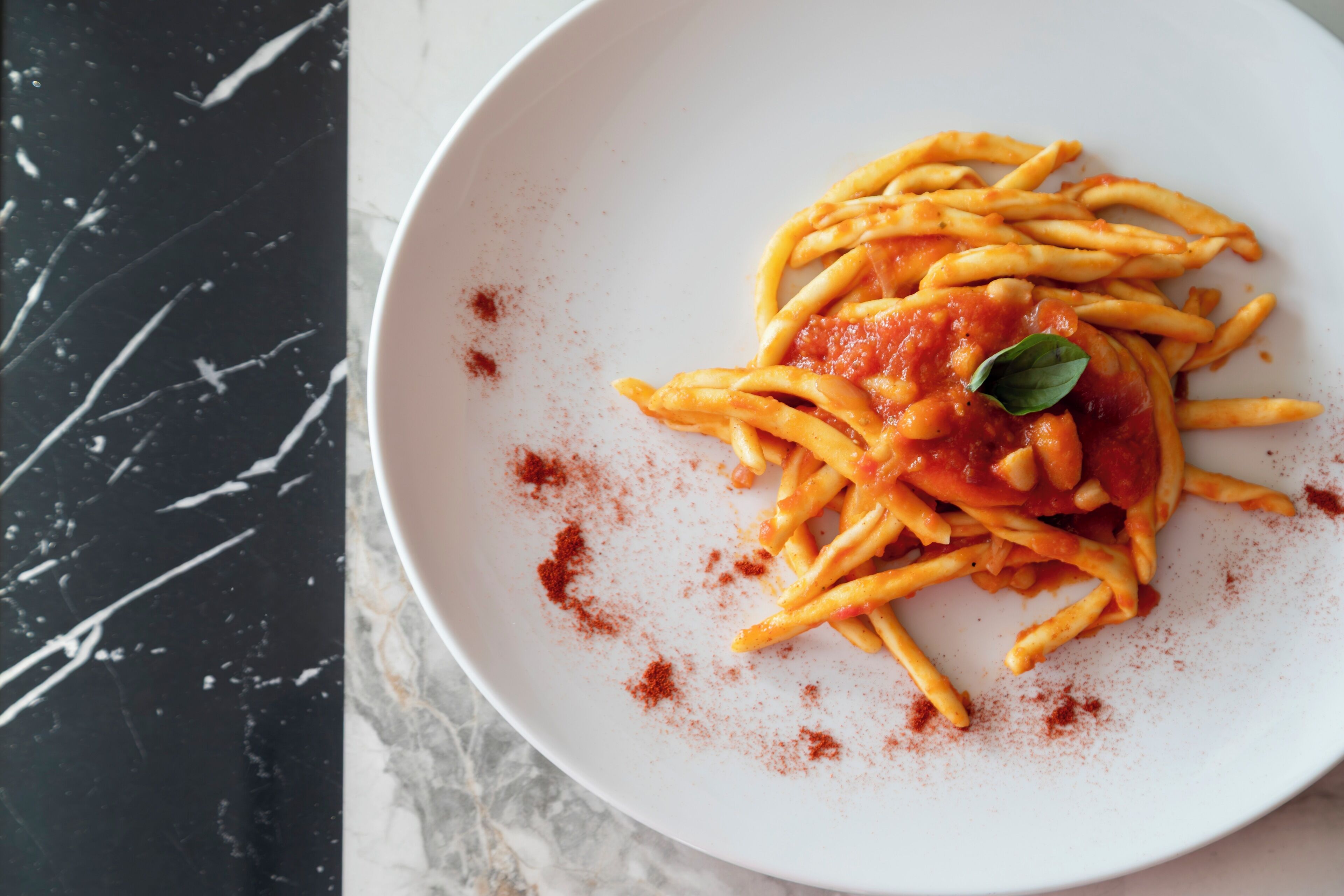 Un plato de espagueti con salsa de tomate y una hoja de albahaca como decoración, espolvoreado con condimento rojo, sobre un plato blanco.