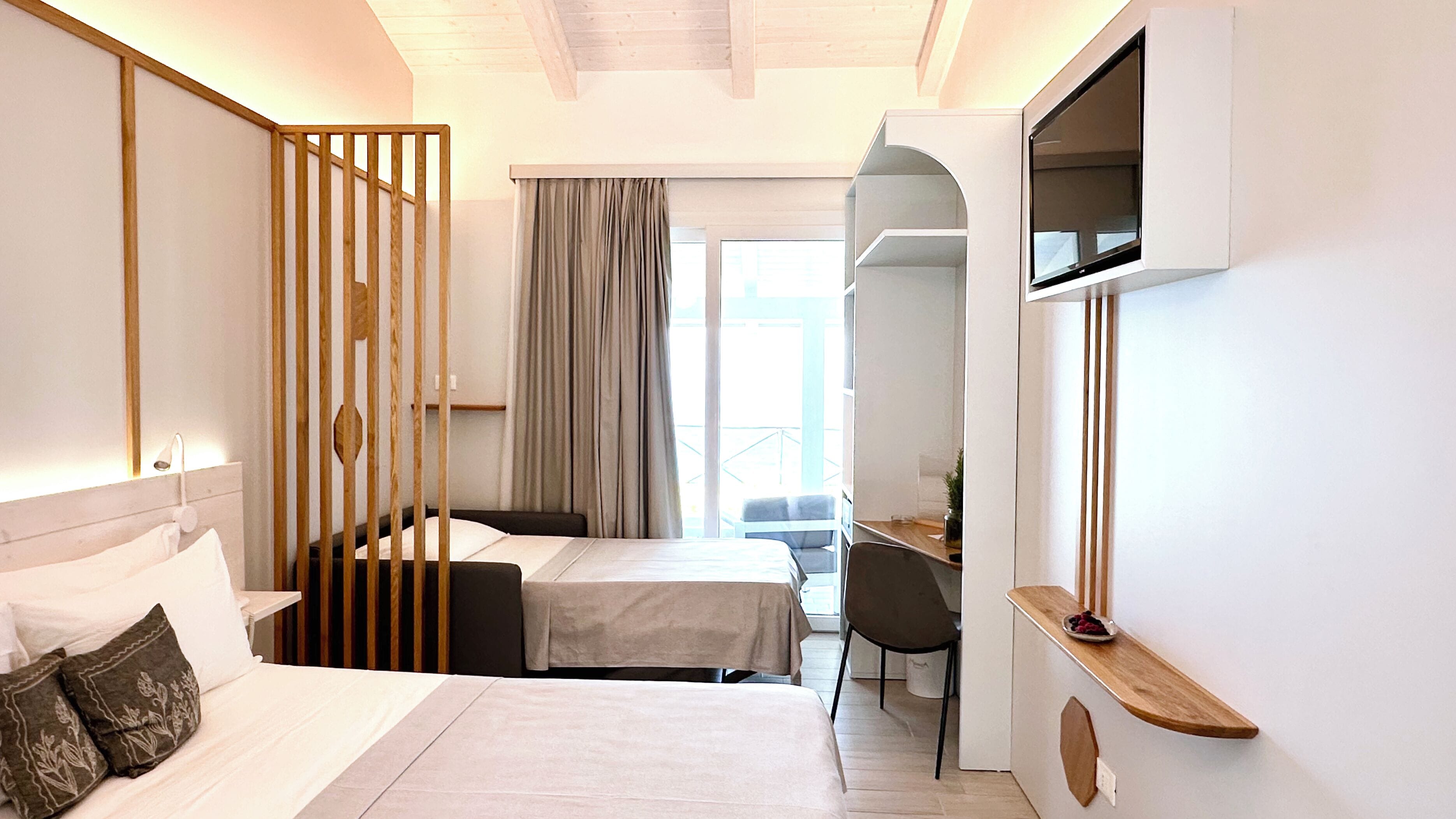 A modern hotel room with a bed, a desk, and a chair. There is a window with curtains, a mirror, and a small table with a bowl of fruit.