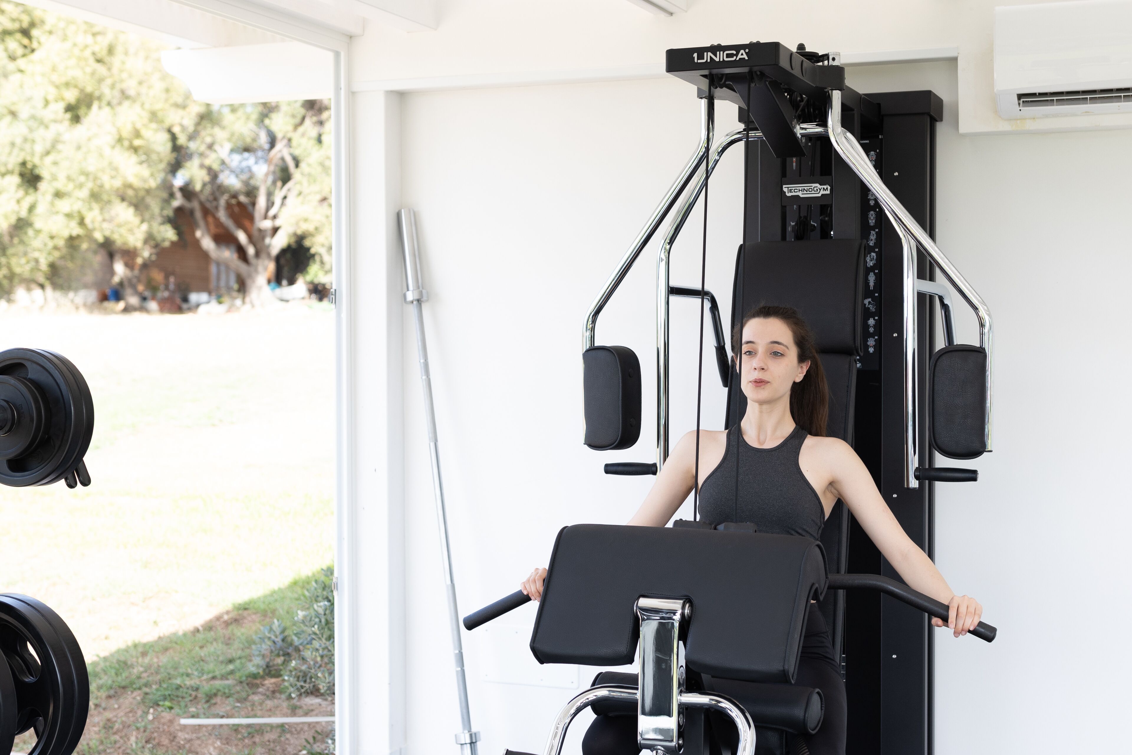 A woman using a weight machine in a gym with a view of trees and a building outside.