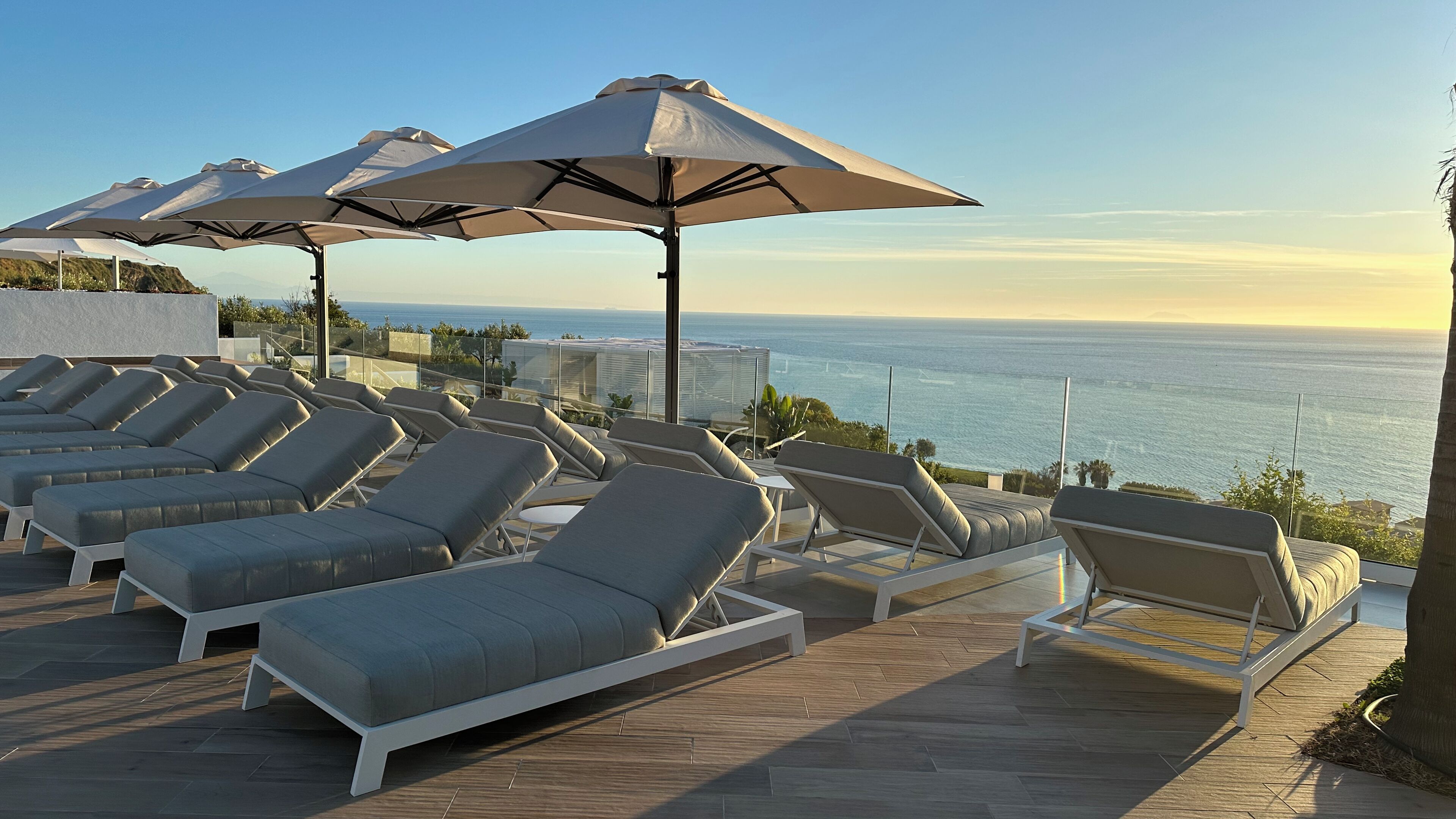 A rooftop lounge area with multiple sun loungers and umbrellas, overlooking a coastal view.