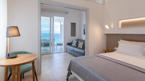A hotel room with a bed, a desk, a chair, a sofa, and a view of the sea through a glass door.