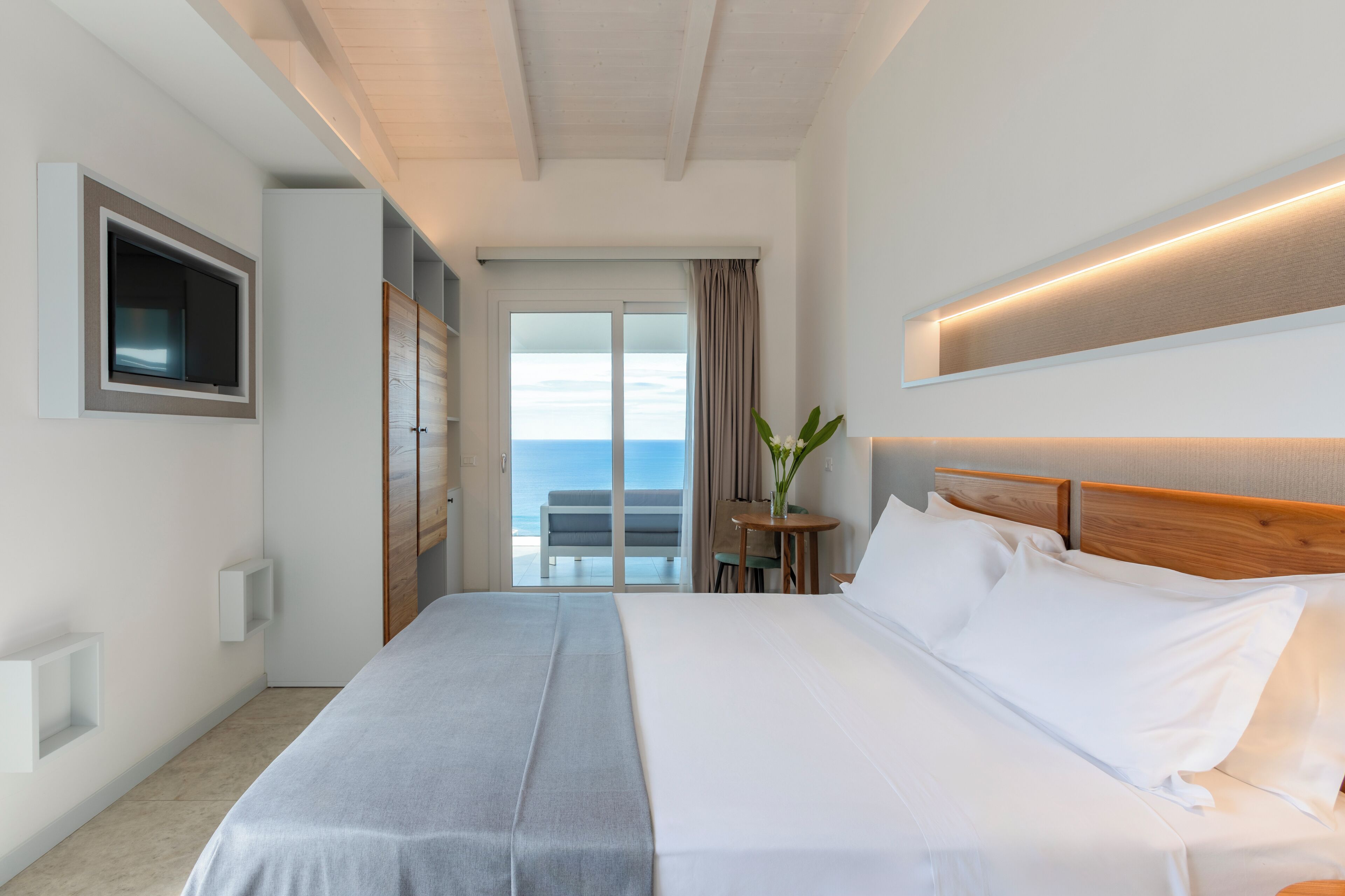 A hotel room with a large bed, a TV mounted on the wall, a small table with a plant, and a view of the ocean through the sliding doors.