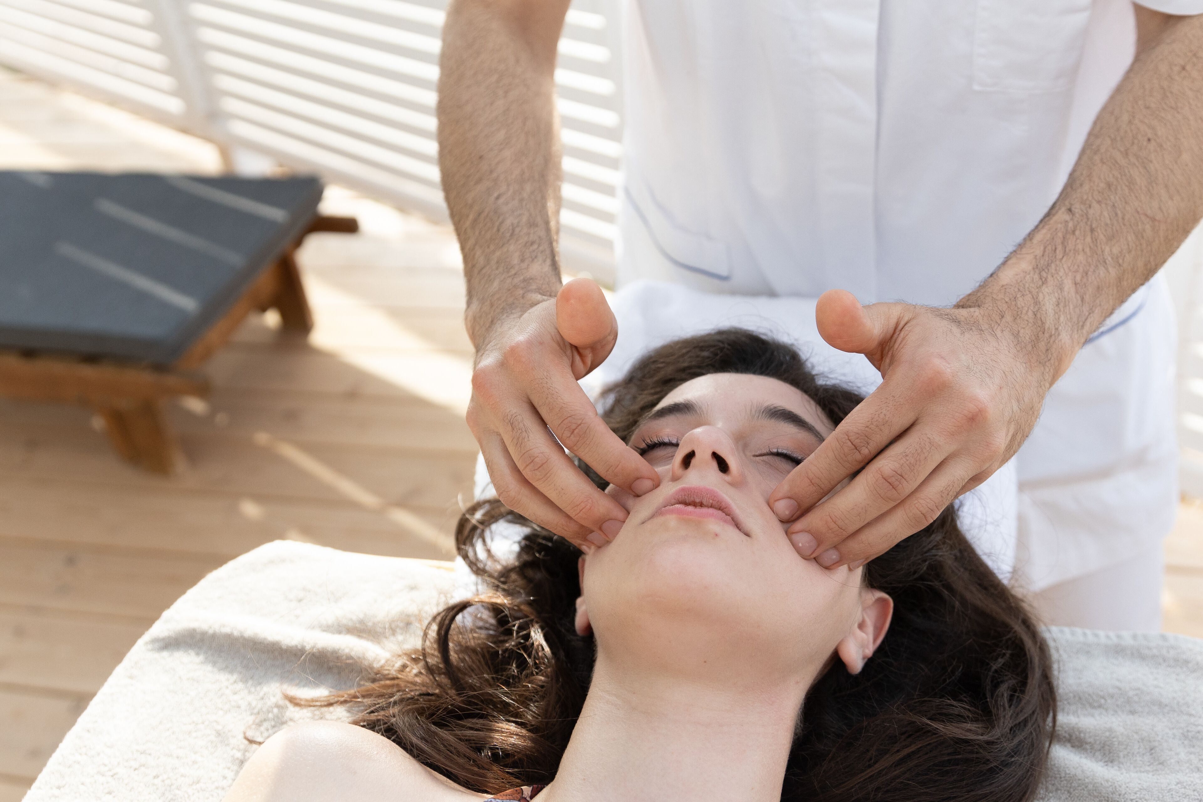 A person receiving a facial treatment in a room with a wooden floor and a relaxation area with a lounge chair.