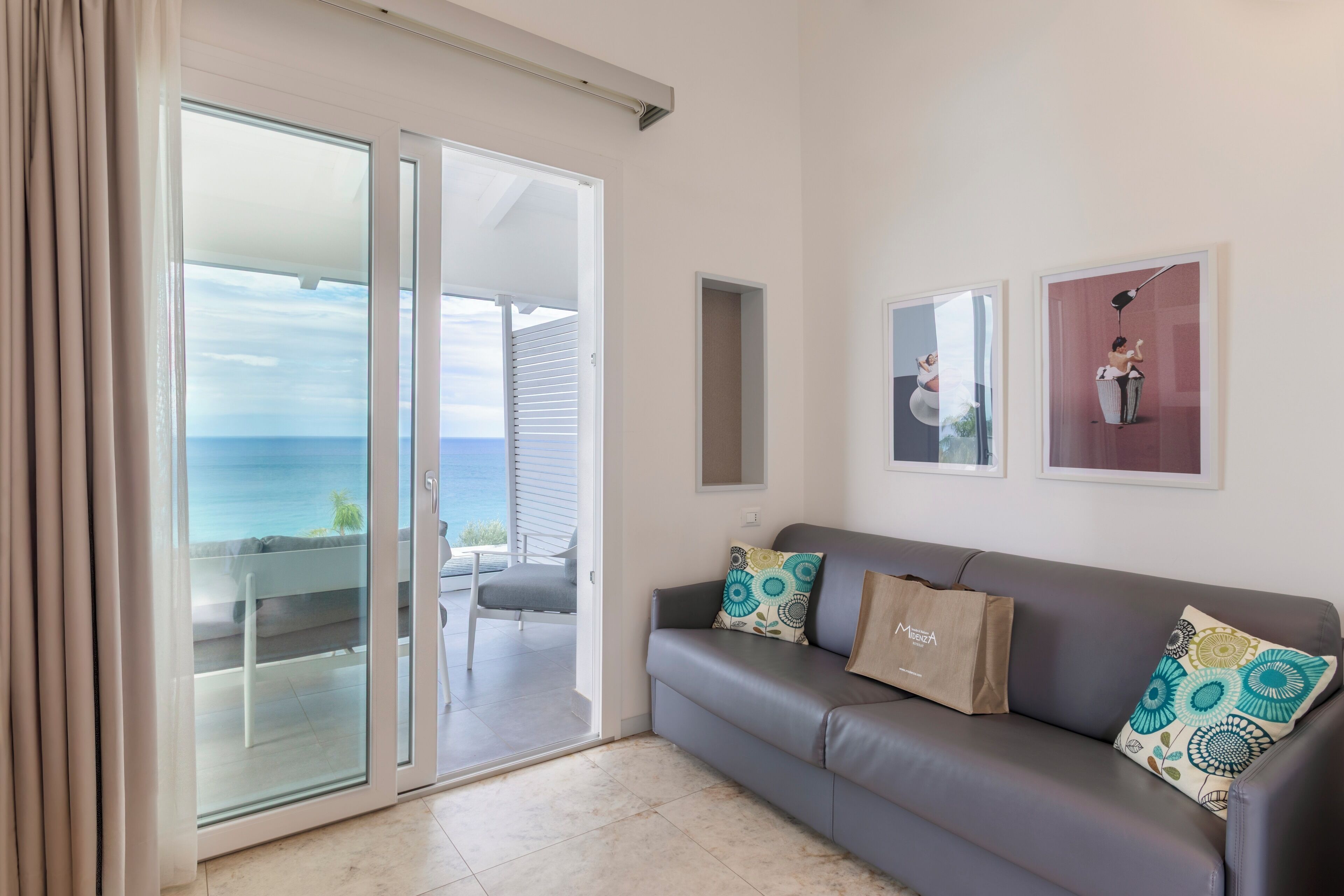 Un salon moderne avec un canapé gris, deux coussins à motifs, un sac en papier marron et une porte en verre donnant sur un balcon avec vue sur la mer.