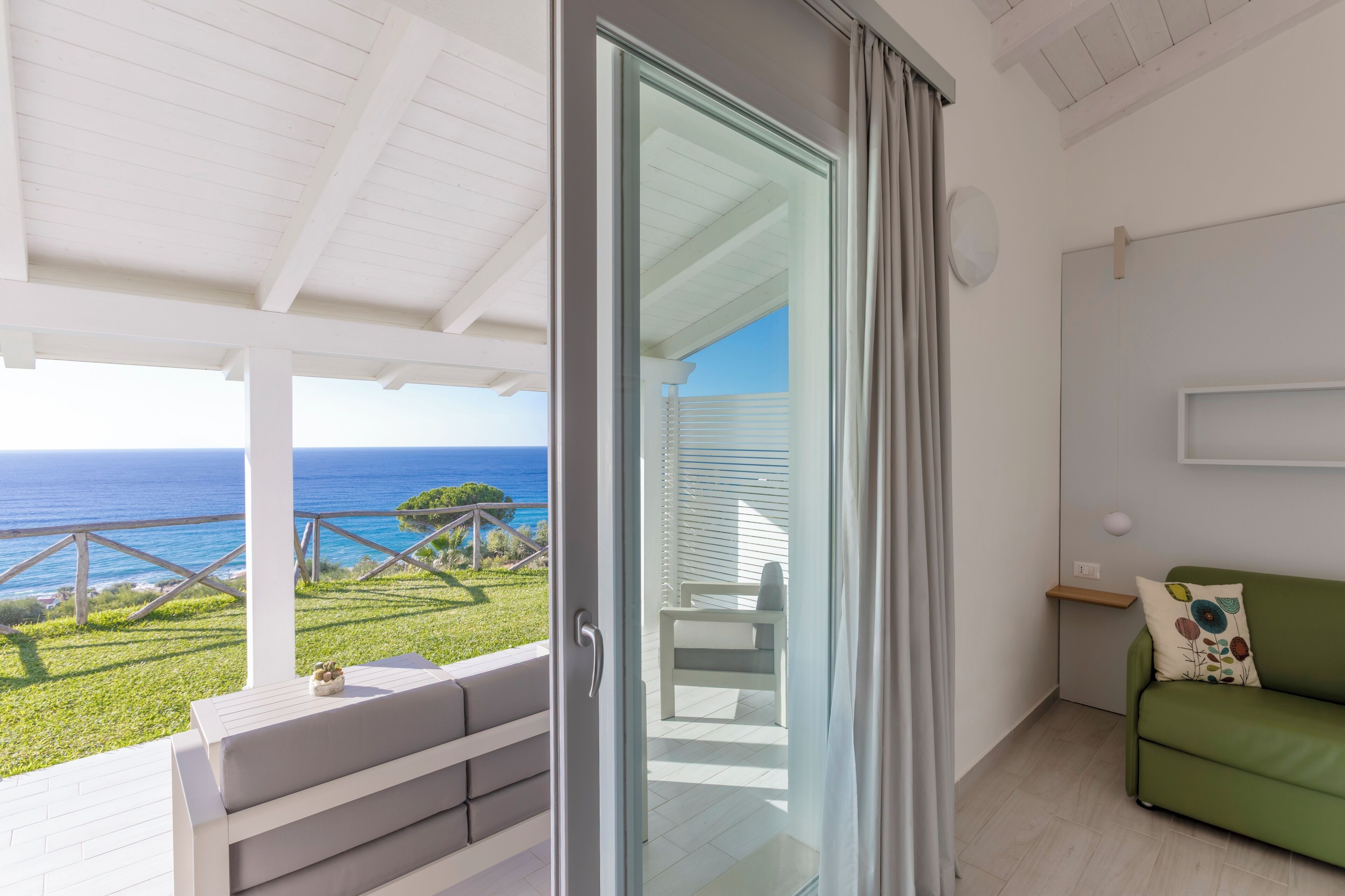 A balcony with a view of the sea, a green sofa, and a sliding glass door leading to a balcony with a chair.
