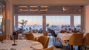 A restaurant with tables set for dining, large windows offering a view of the sea, and a modern ceiling design.