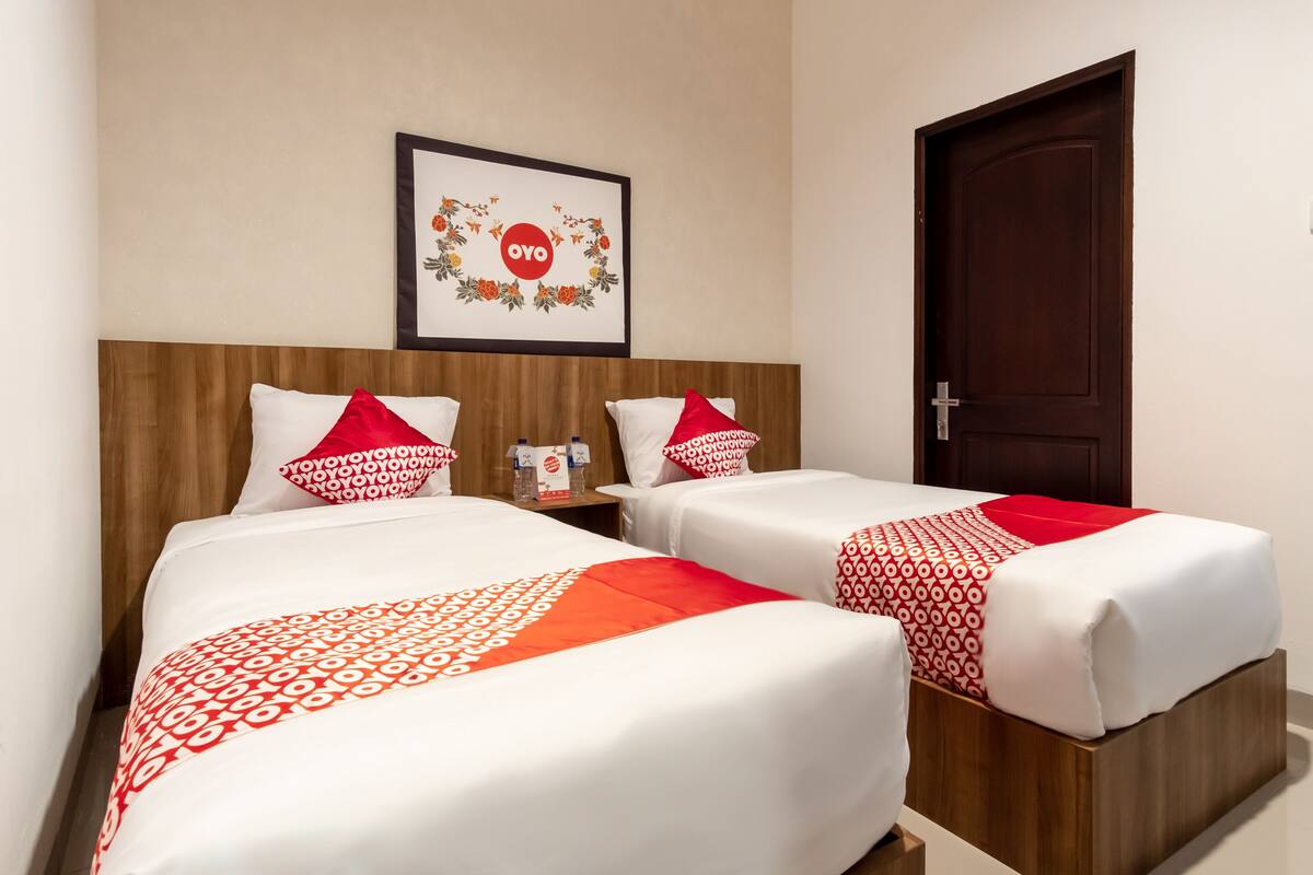 A hotel room with two beds, each with white and red bedding, a framed artwork on the wall, and a wooden door.