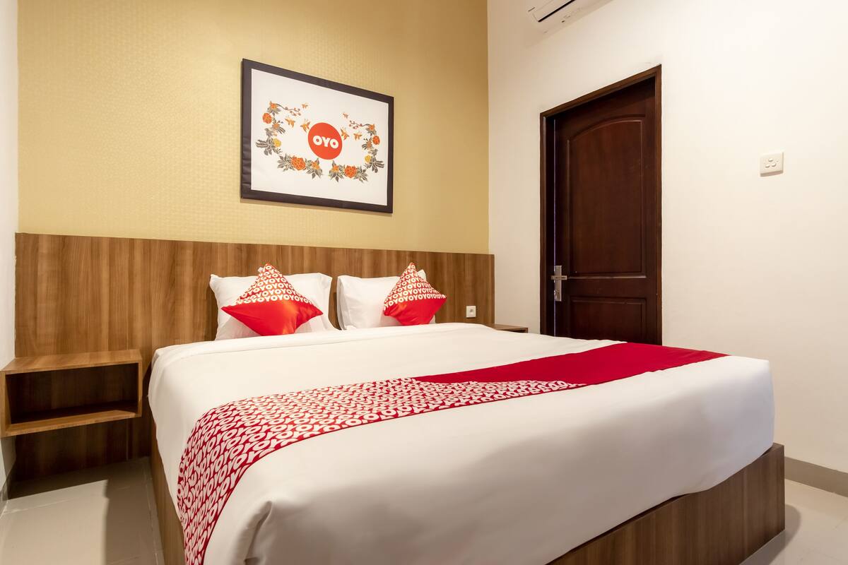 A neatly made bed with a red and white patterned bedspread and pillows, a wooden headboard, a framed picture on the wall, and a closed door.