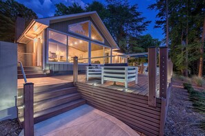 Una casa moderna con terraza de madera, ventanas amplias y una escalera que conduce a la entrada.