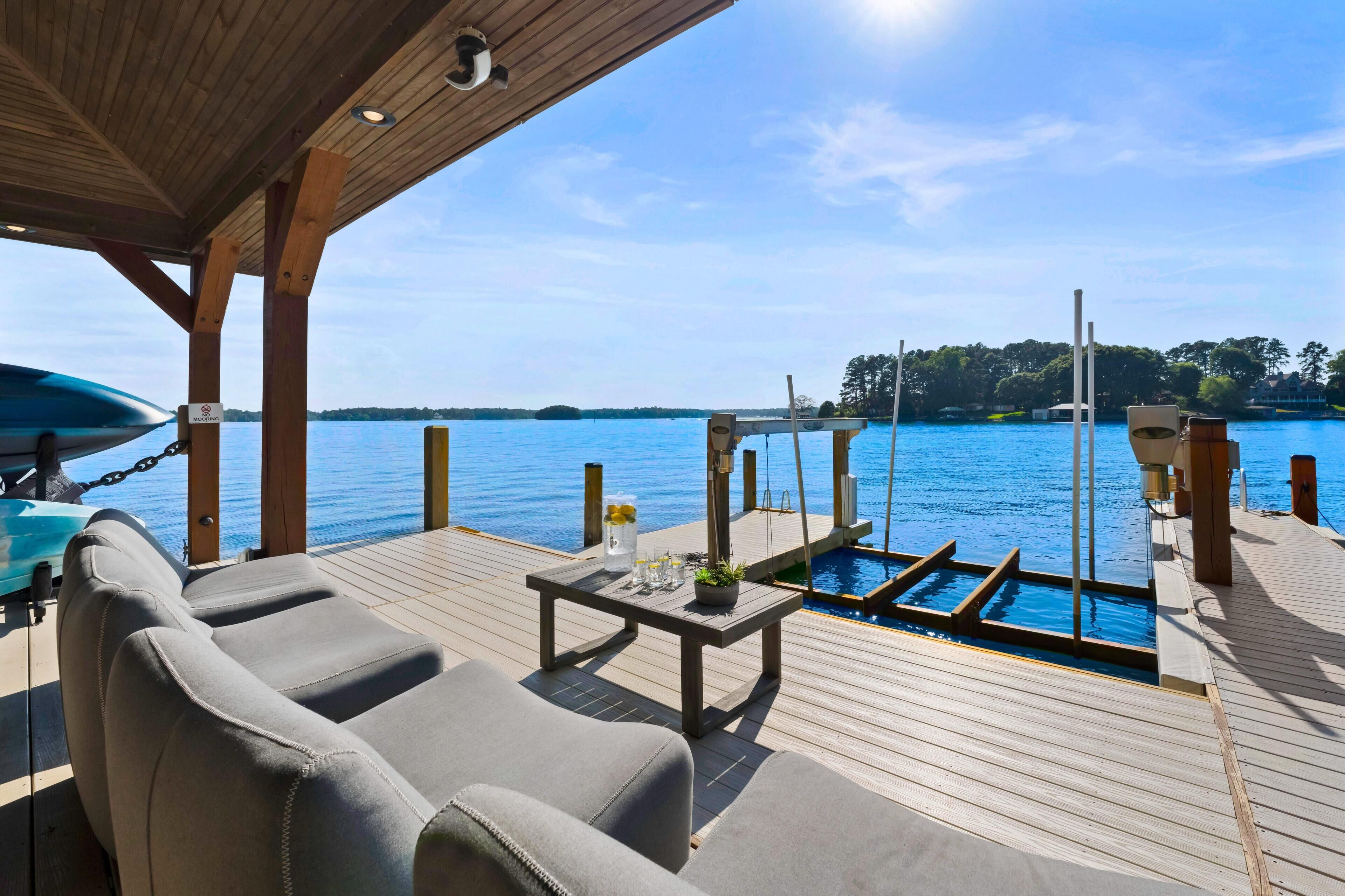 A wooden deck with lounge chairs and a small pool, overlooking a calm body of water with a clear sky.