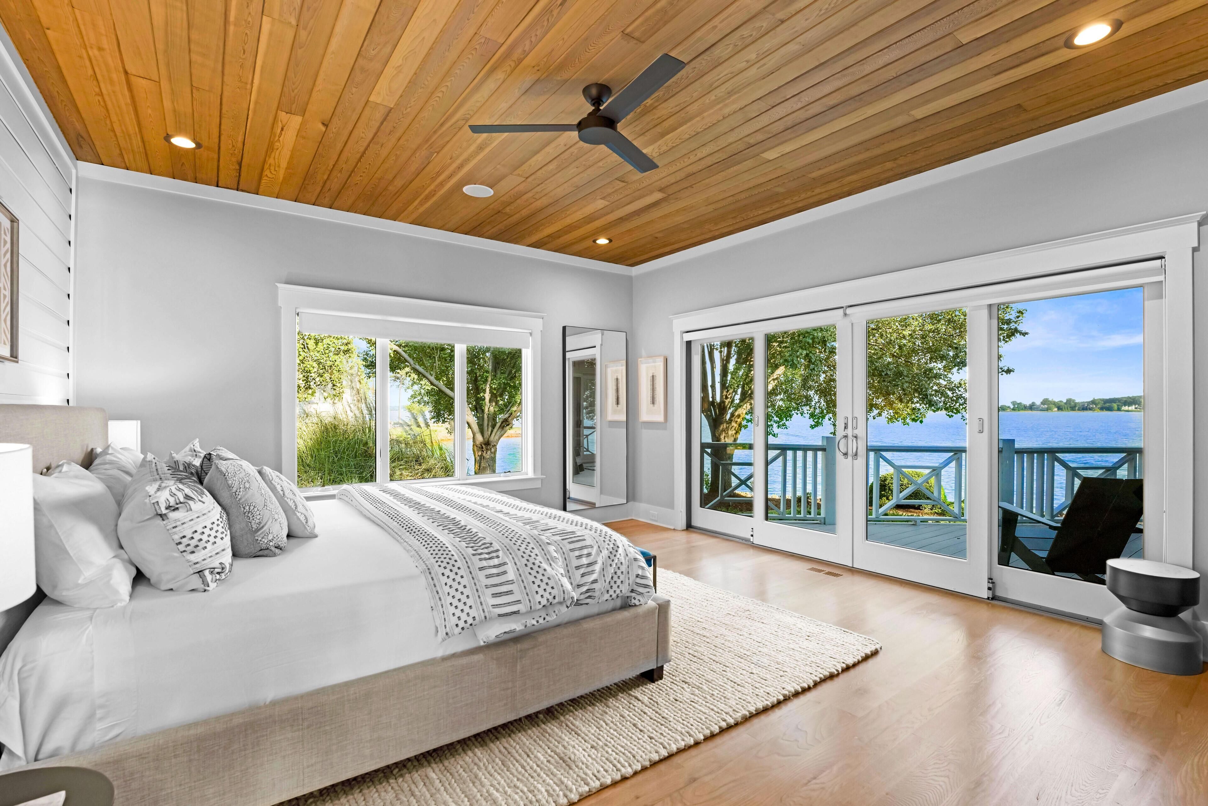 A bedroom with a large bed, a ceiling fan, and a view of a lake through sliding glass doors.