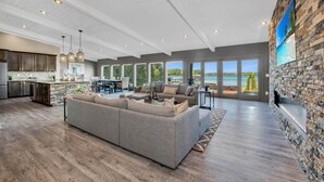 A spacious living area with a large gray sectional sofa, a kitchen with dark wooden cabinets, and a stone accent wall with a TV. - Lake Norman Breakaway (Mooresville)