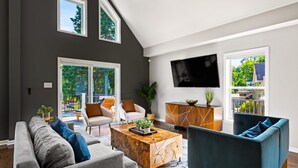 Een moderne woonkamer met een grijze bank, een blauwe fauteuil, een houten salontafel en een aan de muur bevestigde televisie.