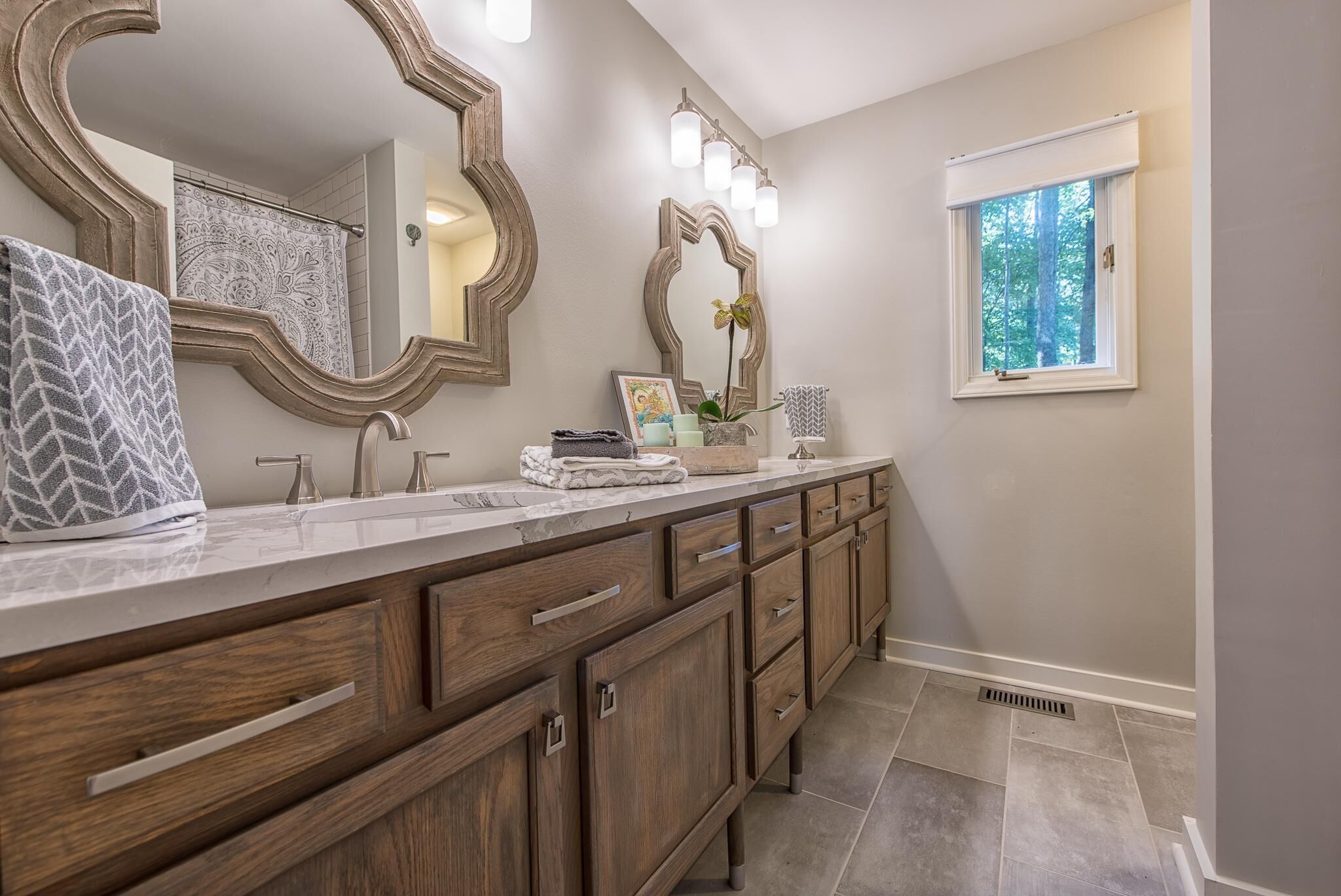 A bathroom with a large vanity, two sinks, wooden cabinets, a large mirror, and a window with a view of trees.