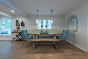 Een eetgedeelte met een houten tafel, twee stoelen, een bank, hangende lampen en een raam met uitzicht op bomen.
