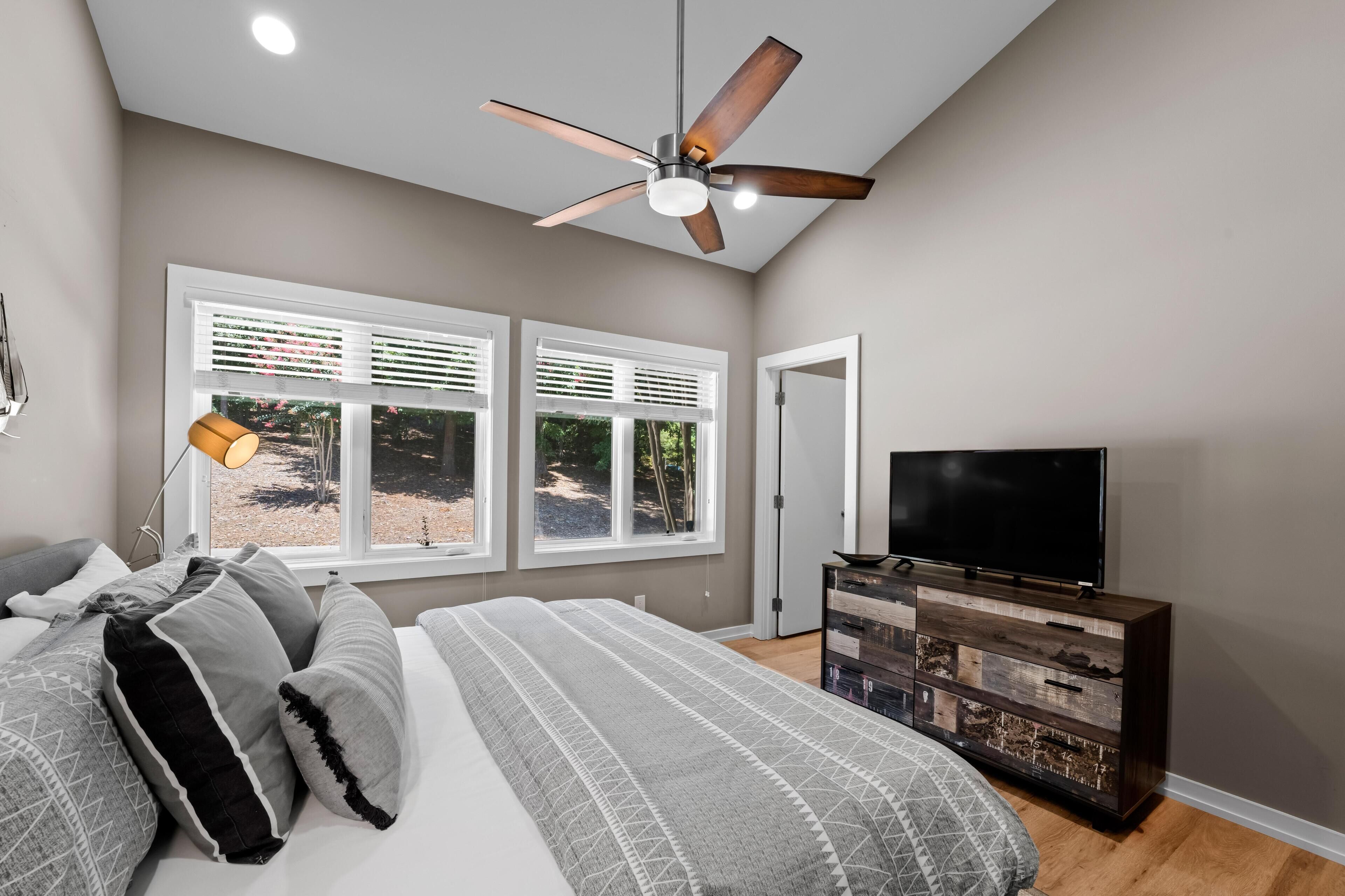 A bedroom with a bed, a ceiling fan, a TV, a dresser, and windows with blinds.