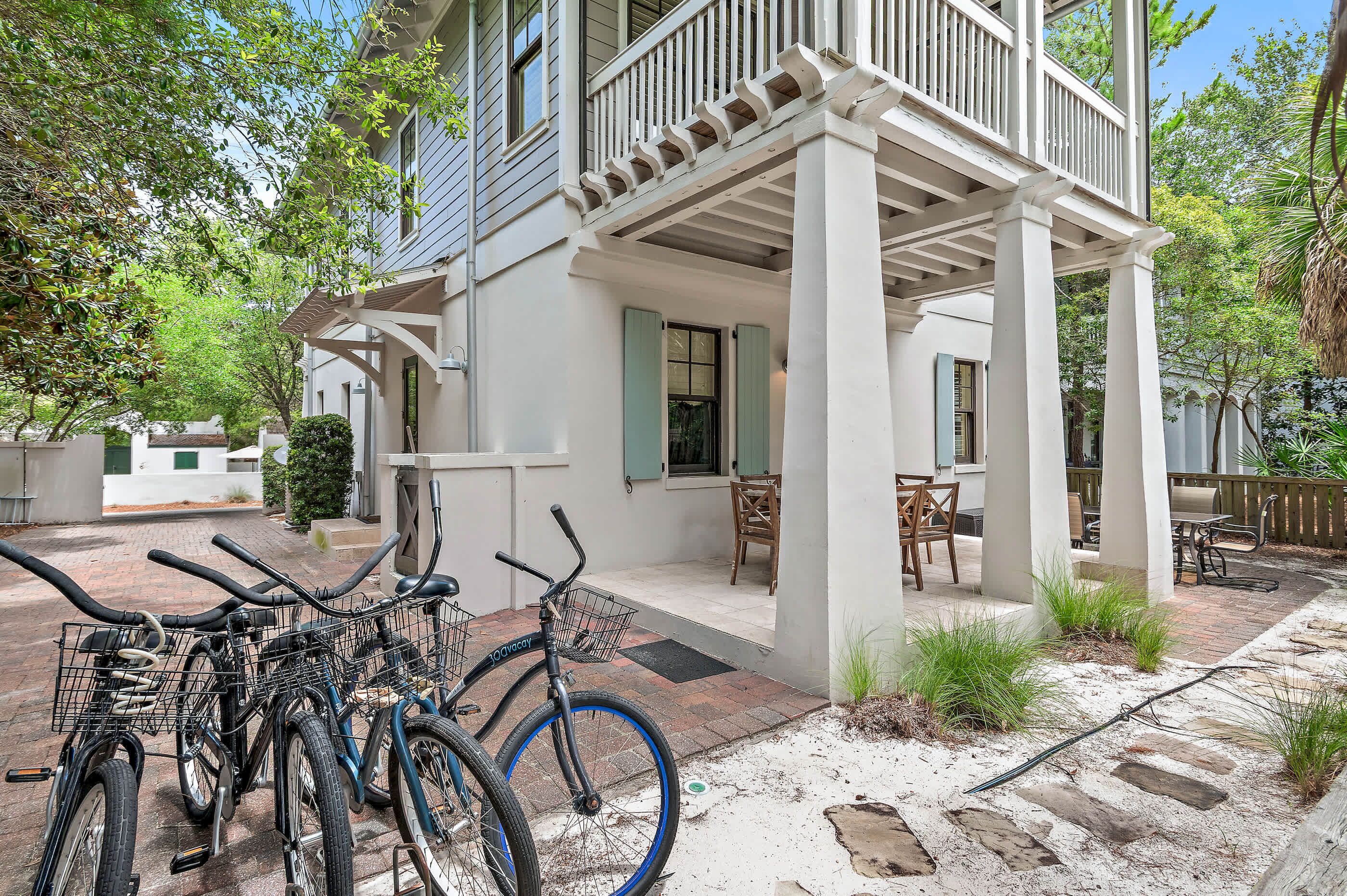 A covered outdoor patio with a brick floor, a white building with green shutters, and a bicycle rack with bicycles.