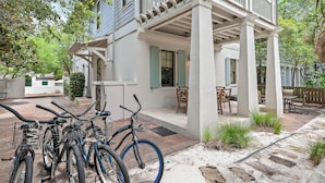 A covered outdoor patio with a brick floor, a white building with green shutters, and a bicycle rack with bicycles.