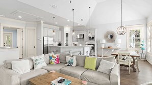 A modern kitchen with white cabinets, a dining area with a wooden table, and a living room with a beige sofa.