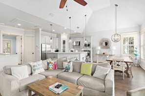 A modern kitchen with white cabinets, a dining area with a wooden table, and a living room with a beige sofa.