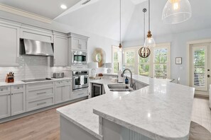 Une cuisine moderne avec des meubles blancs, des appareils en acier inoxydable, un grand îlot central doté d’un évier et des luminaires suspendus.