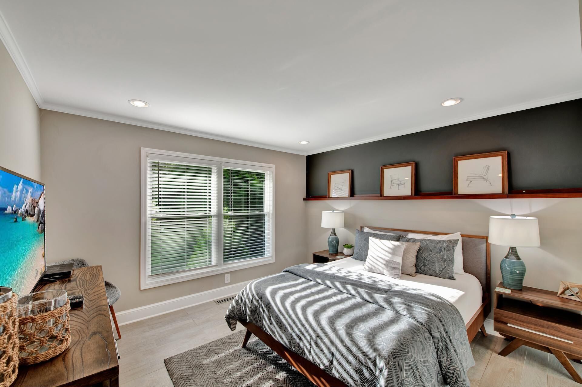 A bedroom with a large bed, two bedside tables with lamps, a window with blinds, and a wall-mounted shelf with framed pictures.