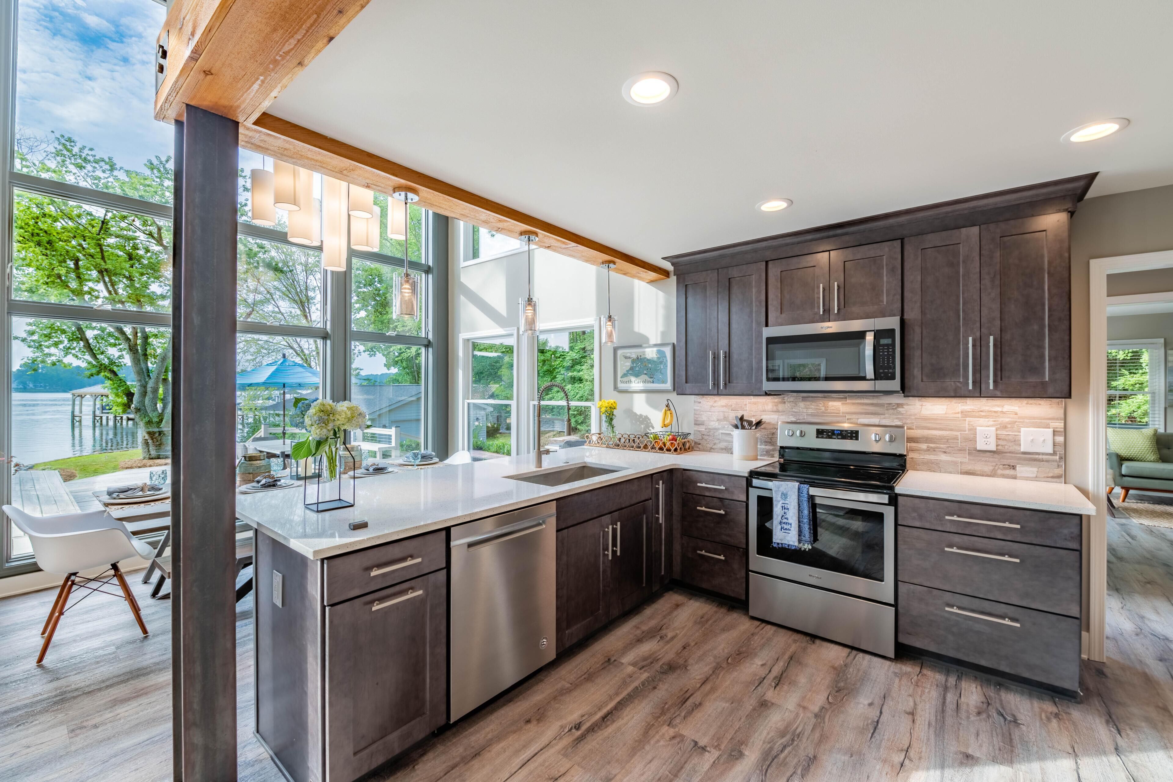 A modern kitchen with dark cabinets, stainless steel appliances, and large windows offering a view of the outdoors.