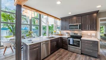 A modern kitchen with dark cabinets, stainless steel appliances, and large windows offering a view of the outdoors.