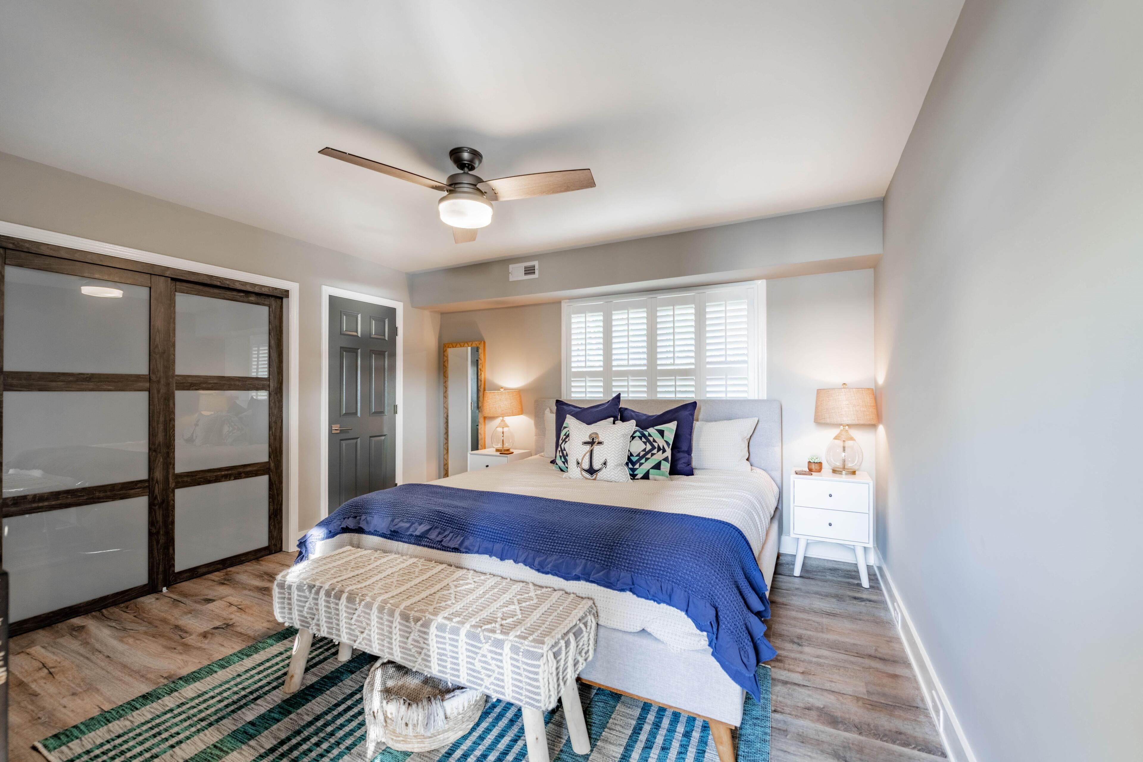 A bedroom with a large bed, a ceiling fan, a window with blinds, and a wooden floor.