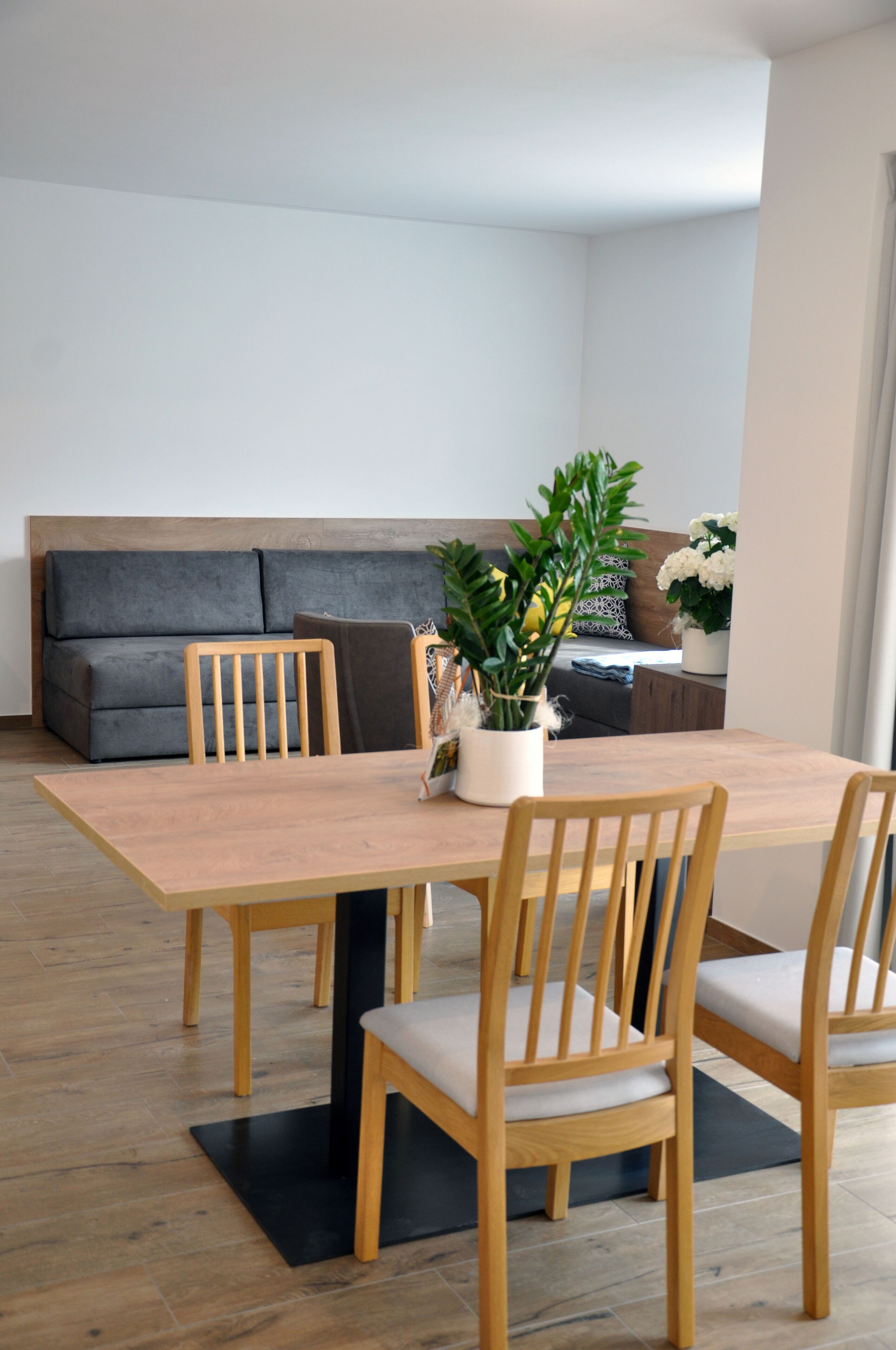 A dining area with a wooden table, four wooden chairs, a grey sofa, and a potted plant.
